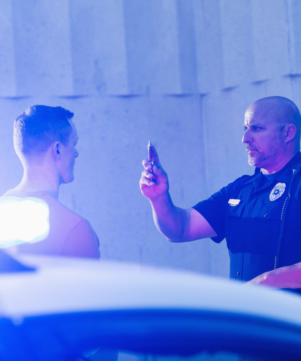 Police officer giving a sobriety test to a young man who he suspects is driving under the influence of drugs or alcohol. Police cruiser is out of focus in the foreground.