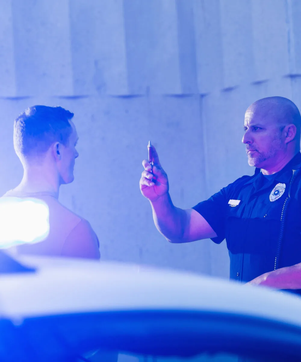 Police officer giving a sobriety test to a young man who he suspects is driving under the influence of drugs or alcohol. Police cruiser is out of focus in the foreground.